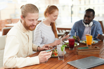 Young bearded man using mobile phone and smiling while have a lunch together with his friends in cafe