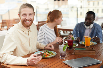 Portrait of bearded young man with mobile phone smiling at camera while have a lunch together with his friends in the background in cafe