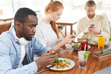 Young people using mobile phones and communicating online while sitting at the table and eating in cafe