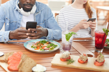 Close-up of African young man sitting at the table and taking a picture of his meal on mobile phone during lunch with his friends