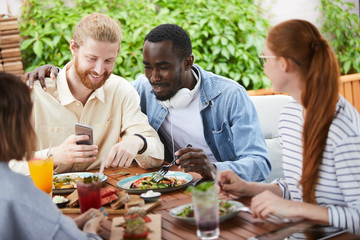 Young man showing some photos on his mobile phone to friends while they sitting at the table and eating during lunch in cafe