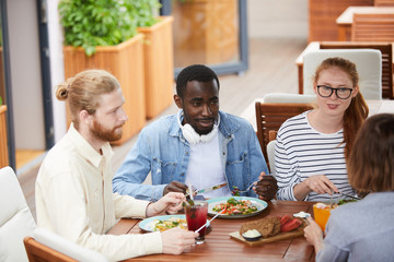 Group of multiethnic young people sitting at the table and talking to each other while have lunch at the restaurant