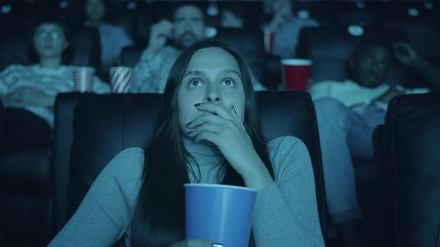 Scared woman watching horror film with open mouth holding popcorn in cinema enjoying interesting movie among men and women. Youth and emotions concept.