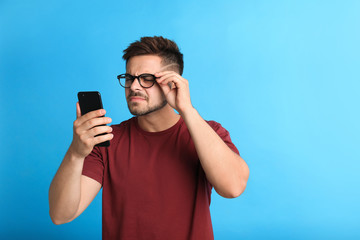 Young man with vision problems using smartphone on blue background, space for text