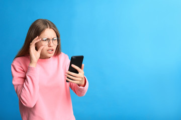 Young woman with vision problems using smartphone on blue background, space for text