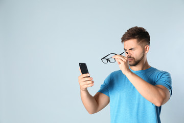Young man with vision problems using smartphone on grey background, space for text