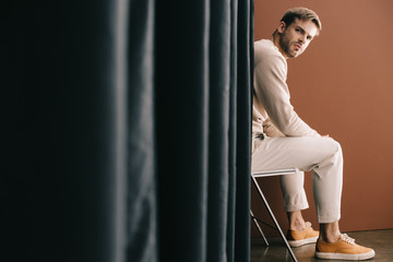 man in casual outfit sitting on chair near curtain on brown