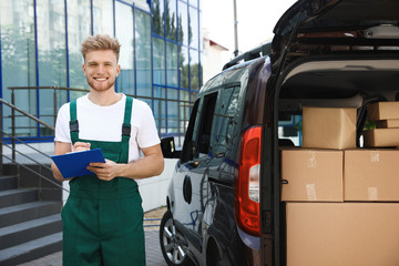 Young courier holding clipboard near delivery van with parcels outdoors