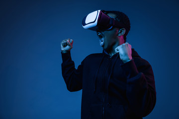 Young african-american man's screaming in VR-glasses in neon on gradient background. Male portrait. Concept of human emotions, facial expression, modern gadgets and technologies. Look like winner.