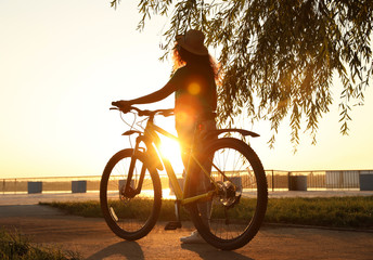 Young African-American woman with bicycle on city waterfront at sunset - obrazy, fototapety, plakaty
