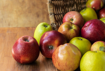 Red and green apples spilled out from a wicker basket lying on its side onto a brown wooden board, side view