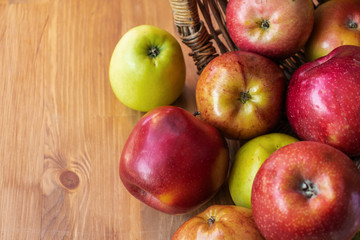 Red, green and yellow apples rolled out of a wicker basket onto a brown wooden surface, top side view