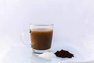 A fresh cup of coffee in a glass mug. There is s small pile of sugar and coffee in front