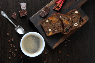 Black coffee and dark bread with nuts and dried fruits. On the surface of the table, pieces of dark chocolate and coffee beans are laid out. Dark background. View from above.
