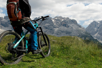 Fototapeta premium man with electric bike, e-bike, ebike, looking mountains of Tosa and Brenta Peak, Grosté Pass, meadow, Dolomites, Madonna di Campiglio, summer, sport, adventure, travel, Alps, Trentino, Italy