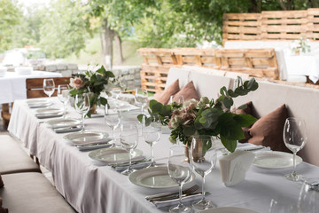 Wedding table setting decorated with fresh flowers in a brass vase. Wedding floristry. Banquet table for guests outdoors with a view of green nature. Bouquet with roses, eustoma and eucalyptus leaves