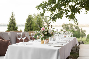 Wedding table setting decorated with fresh flowers in a brass vase. Wedding floristry. Banquet table for guests outdoors with a view of green nature. Bouquet with roses, eustoma and eucalyptus leaves