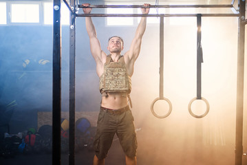 Young sports man with heavy waistcoat makes pull ups on crossbar in the gym full of smoke.