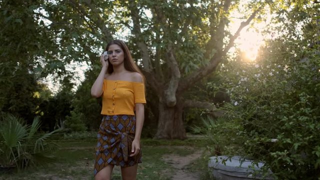Medium shot of beautiful latin girl passing by putting flower in hair dreamily looking in camera in old garden at sunset
