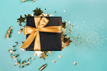 Black gift box with golden bow on turquoise background with glitter. Holiday concept.
