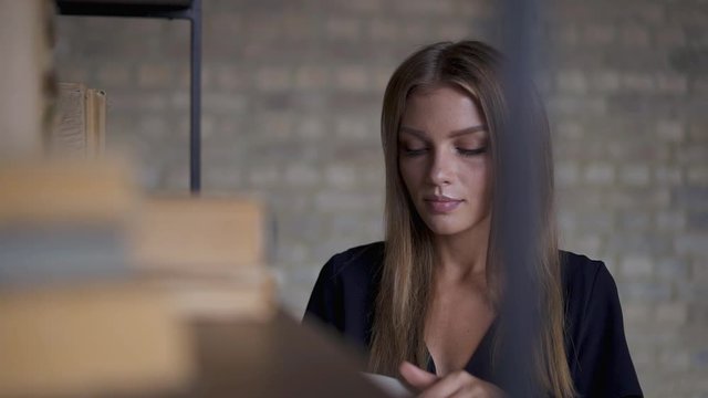 Beautiful young caucasian woman with long hair wearing black dress taking book from shelf, reading and hugging it with smile. Concept of favourite book. Slider slow motion medium shot