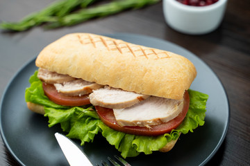 Close up of toasted ciabatta with tomatoes, smoked chicken breast and bunch of green leaves with olive oil and pepper, served on dark wood table with knife and fork