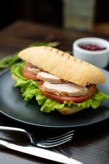 Close up of toasted ciabatta with tomatoes, smoked chicken breast and bunch of green leaves with olive oil and pepper, served on dark wood table with knife and fork
