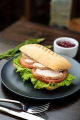 Close up of toasted ciabatta with tomatoes, smoked chicken breast and bunch of green leaves with olive oil and pepper, served on dark wood table with knife and fork