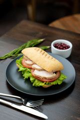 Close up of toasted ciabatta with tomatoes, smoked chicken breast and bunch of green leaves with olive oil and pepper, served on dark wood table with knife and fork