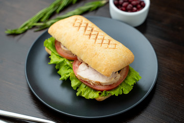 Close up of toasted ciabatta with tomatoes, smoked chicken breast and bunch of green leaves with olive oil and pepper, served on dark wood table with knife and fork