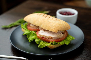 Close up of toasted ciabatta with tomatoes, smoked chicken breast and bunch of green leaves with olive oil and pepper, served on dark wood table with knife and fork