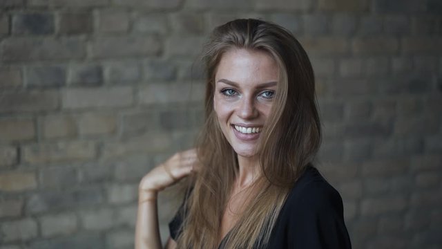 Beautiful blonde young woman in black dress smiling, playing with her hair, flirting and winking, then going away. Handheld slow motion medium shot