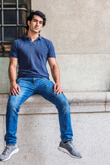 Portrait of Young Mix Race American College Student in New York City, wearing blue short sleeve shirt, blue jeans, sneakers, sitting against old style wall on street in campus, relaxing, thinking..