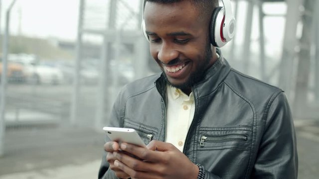 Happy African American student in headphones touching smartphone screen outdoors smiling enjoying multimedia content standing alone in city street.