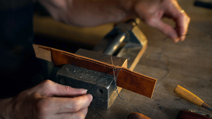 Man sews leather details clamped in vice. Craftsman works with needle and thread. Small business concept.