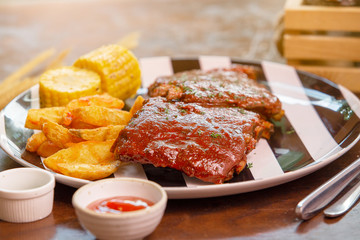 Spicy hot grilled spare ribs from a summer BBQ served with a grilled potato wedges and piece of sweet corn on black and white plate on wooden table.
