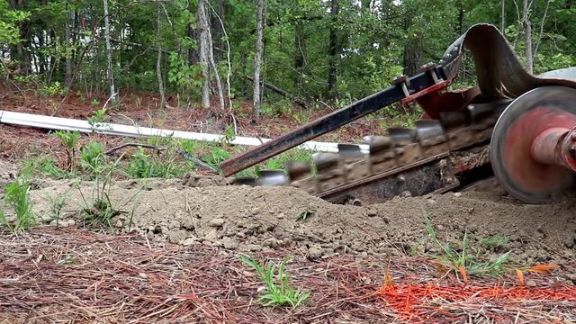 Trenching machine cuts a ditch for backyard irrigation with large blade slicing through the soil and auger spinning. Utility trench being cut in sandy soil.