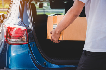 Delivery man is delivering cardboard box to customers via private car trunk door. People lifestyles and business occupation concept. Young male courier in casual clothes. Parcel move courier service