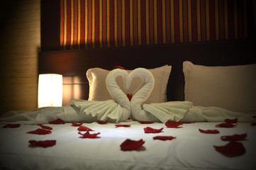 Cama de hotel con pétalos de rosas y toallas con forma de cisnes creando un corazón. Noche...