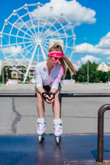 Portrait of an emotional girl in a pink cap visor wearing protective gloves and rollerblades sitting on the background of ferris wheel.