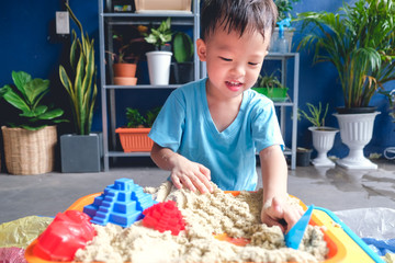Cute smiling Asian 3 - 4 years old toddler boy playing with kinetic sand in sandbox at home /...
