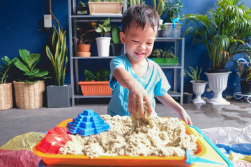 Cute smiling Asian 3 - 4 years old toddler boy playing with kinetic sand in sandbox at home /...