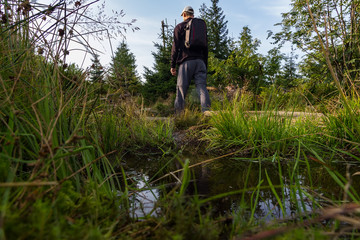 Naklejka na ściany i meble Young man tourist in white cap with backpack walking near small pond in landscape with trees, Sumava National Park and Bavarian Forest, Czech republic and Germany