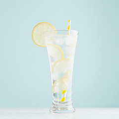 Cold bright organic homemade cocktail with lemon slices, ice cubes, tonic, yellow in light soft pastel blue  background on white wood board, square, closeup.
