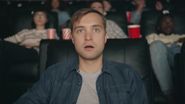 Emotional young man student is watching shocking film opening mouth touching fece with hand in dark cinema. Emotions, youth and entertainment concept.
