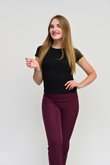 Full-length vertical portrait of a pretty brunette girl with long flowing hair in a black t-shirt and burgundy trousers on a white background. Smiling, showing emotions.