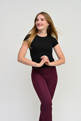 Naklejka premium Full-length vertical portrait of a pretty brunette girl with long flowing hair in a black t-shirt and burgundy trousers on a white background. Smiling, showing emotions.