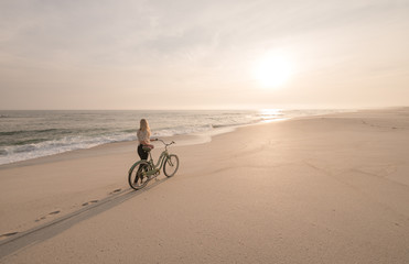 Beautiful blonde woman and bicycle traveling down a quiet beach at sunset.