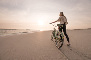 Beautiful blonde woman and bicycle traveling down a quiet beach at sunset.