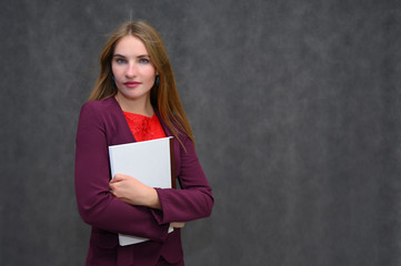Portrait of a pretty secretary manager brunette girl with long flying hair in a burgundy business suit with a folder on a gray background. Smiling, showing emotions.